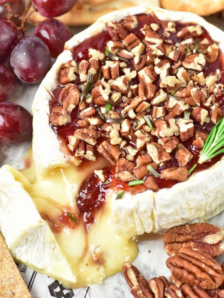 Baked Brie topped with red jam, chopped pecans, and rosemary sprigs, with grapes and crackers nearby.