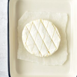 A whole wheel of brie scored with a diamond pattern on parchment paper in a baking dish.