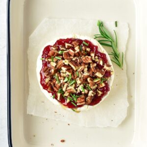 Round wheel of brie topped with cranberry sauce, chopped pecans, and rosemary, served on parchment paper.