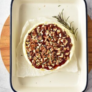 A wheel of baked brie topped with jam, chopped pecans, and a sprig of rosemary in a baking dish.