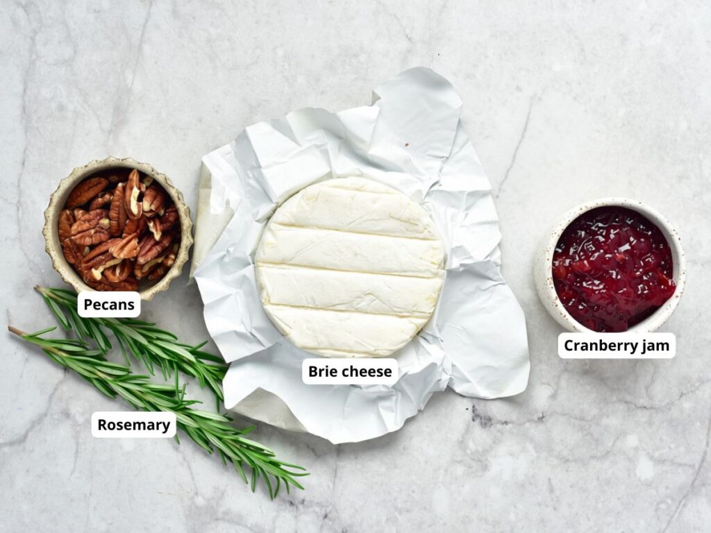 A round of brie cheese in white paper, a bowl of pecans, a bowl of cranberry jam, and sprigs of rosemary, with labels identifying each item.