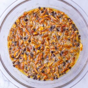 A glass bowl containing a mixture of pecans, chocolate chips, and melted butter.