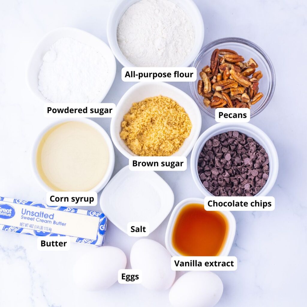 Baking ingredients arranged on a white surface, labeled: all-purpose flour, pecans, powdered sugar, brown sugar, chocolate chips, corn syrup, salt, vanilla extract, butter, and eggs.