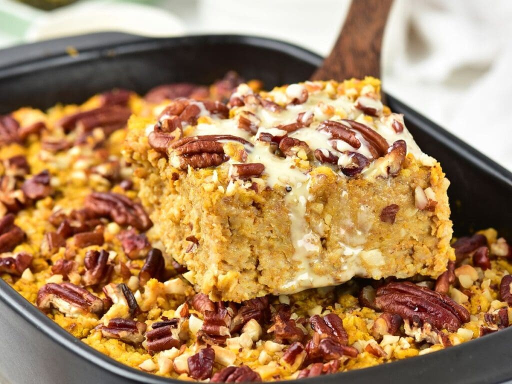 A slice of pumpkin casserole topped with pecans and a cream glaze being lifted from a baking dish with a wooden spatula.