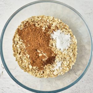 A glass bowl filled with rolled oats, topped with piles of cinnamon, baking powder, and salt.