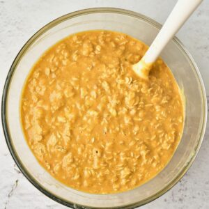 A glass bowl containing orange-tinted oat batter, with a white spatula resting inside.
