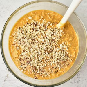 A glass bowl filled with orange batter topped with chopped nuts, with a white spatula resting inside.
