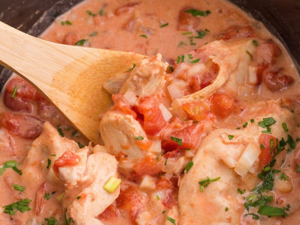 A wooden spoon lifting a piece of chicken from a creamy tomato sauce with diced onions and herbs.