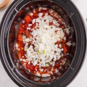 A slow cooker bowl filled with diced tomatoes, chopped white onions, and spices.