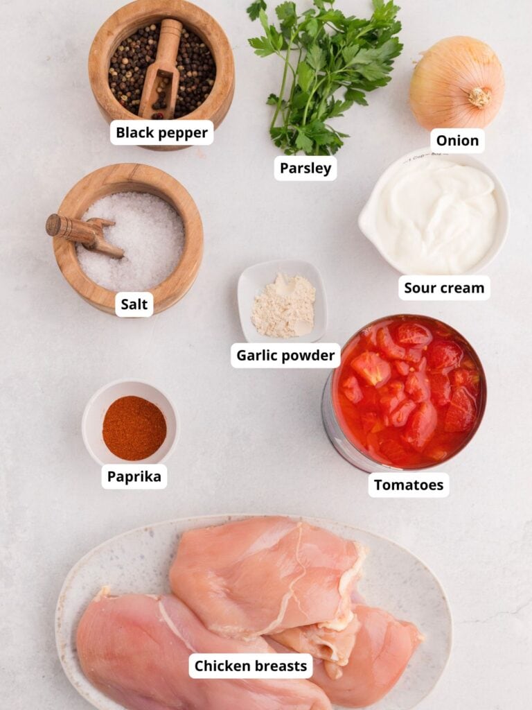 Ingredients for a recipe arranged on a white surface, labeled: black pepper, parsley, onion, salt, sour cream, garlic powder, paprika, canned tomatoes, and raw chicken breasts.