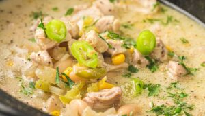 A close-up of white chicken chili containing shredded chicken, white beans, corn, and sliced green peppers.