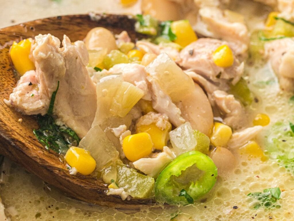 A wooden spoon holding chicken chili with corn, beans, diced onions, and jalapeño slices.