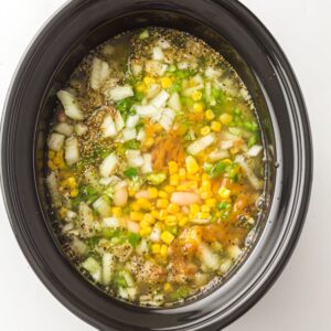 A slow cooker filled with broth, corn, chopped onions, peppers, beans, and spices.