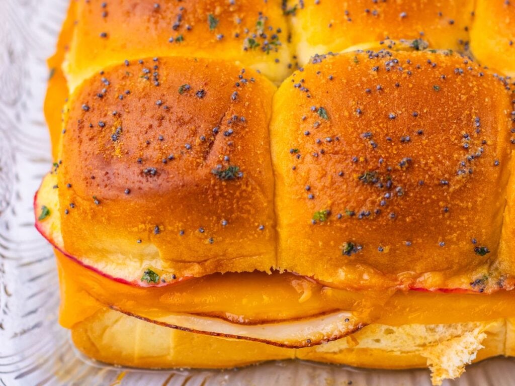 Sliders topped with poppy seeds and herbs, filled with ham and melted cheese.