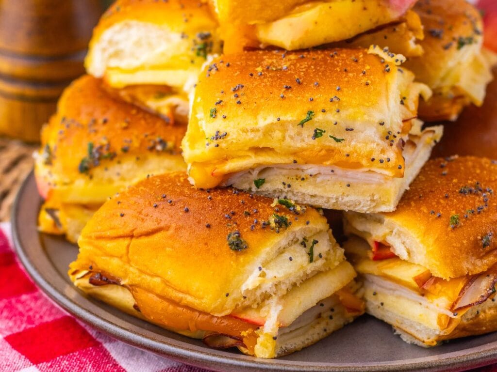 A pile of ham and cheese sliders on a gray plate, topped with poppy seeds and herbs.