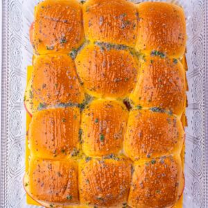 A rectangular tray of twelve baked ham and cheese sliders topped with poppy seeds and herbs.