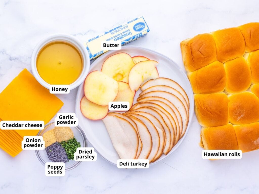 Ingredients for sandwiches laid out on a white surface: sliced apples, deli turkey, Hawaiian rolls, cheddar cheese, a stick of butter, honey, and a small dish containing garlic powder, onion powder, poppy seeds, and parsley.